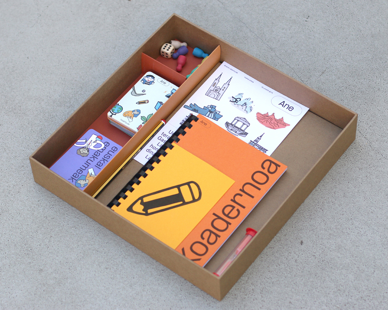 Caja del juego abierta, en la que se ven, las tarjettas con los retos, las fichas, los dados, un rreloj de arena, el tablero y un cuaderno.